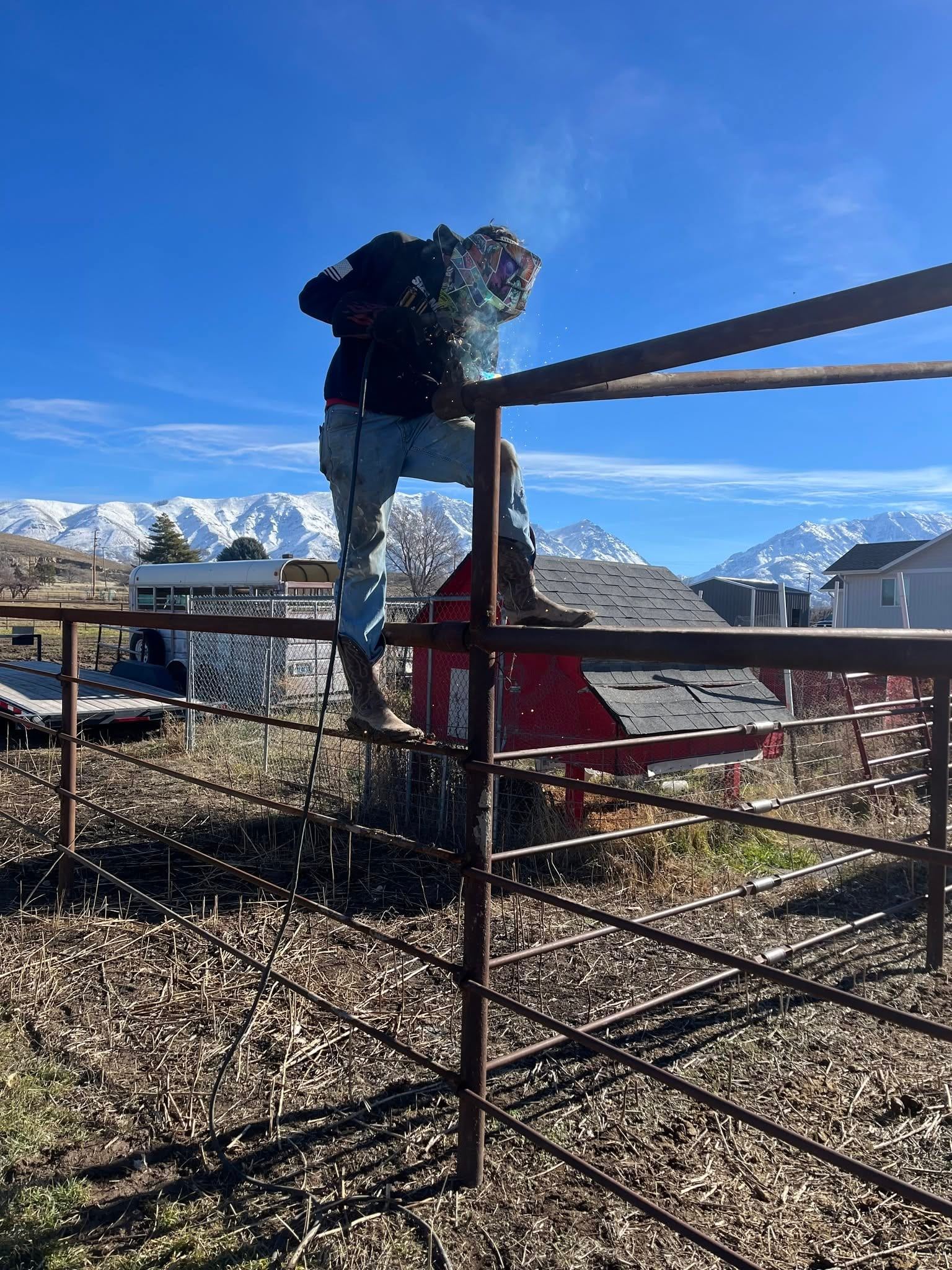 A welder perched on a corral fence, arc lit, with the snow-capped Wasatch range behind