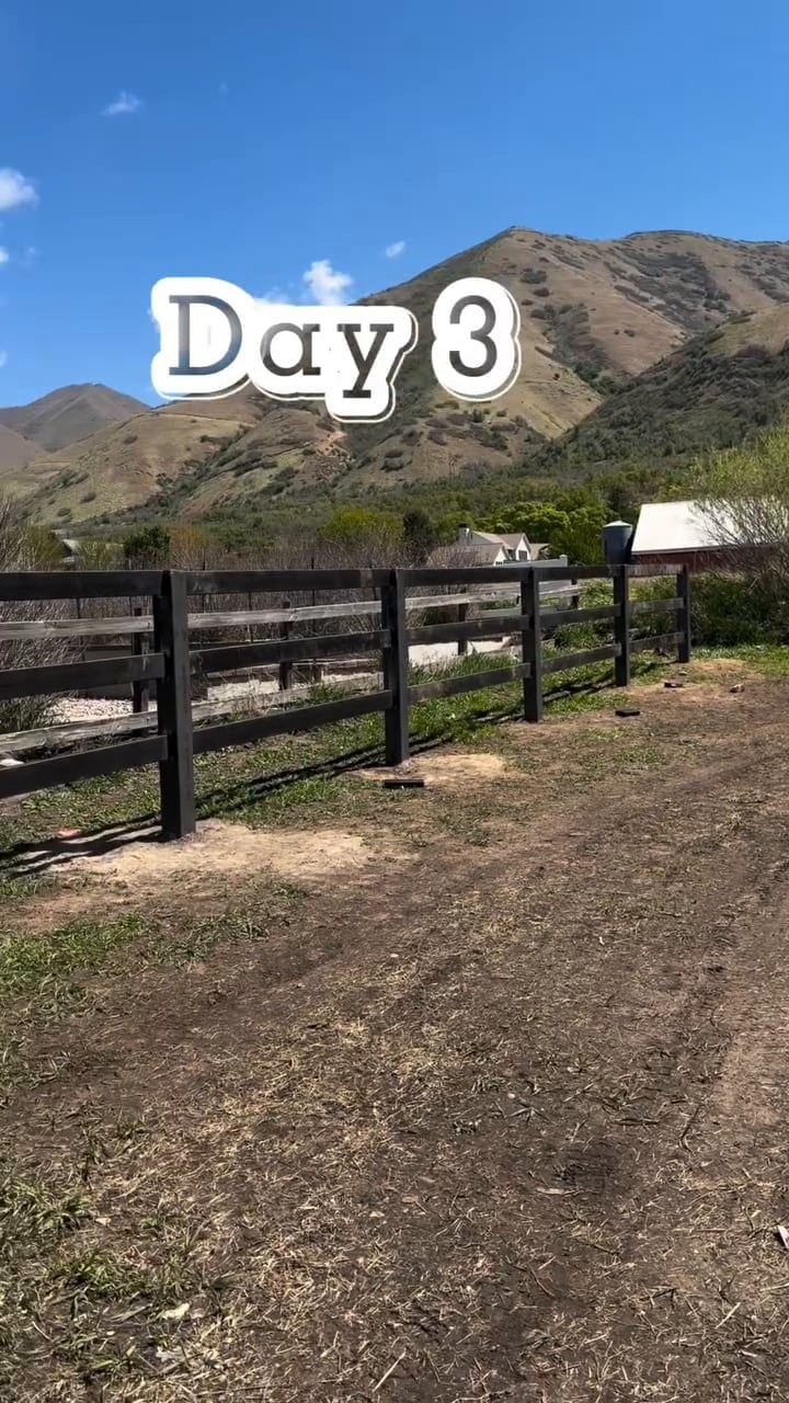 A black ranch fence running across a green field toward the Utah foothills